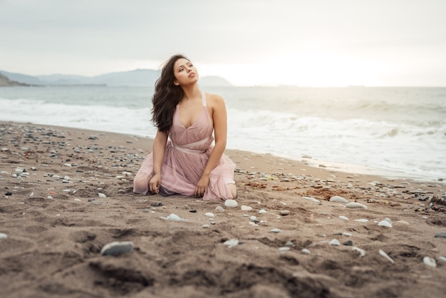 Bella ragazza latina con il lungo bastone nero in posa sulle sue ginocchia su un bel tramonto sulla spiaggia vestita con un abito da balletto rosa