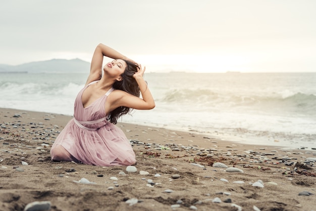 Bella ragazza latina con il lungo bastone nero in posa sulle sue ginocchia su un bel tramonto sulla spiaggia vestita con un abito da balletto rosa