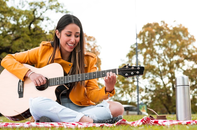 Bella ragazza latina, facendo un picnic e suonando la chitarra in un parco, bevendo compagno, con il cielo sullo sfondo. stile di vita