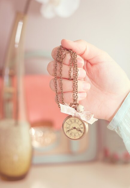 Bella ragazza mano che tiene una piccola collana orologio vintage davanti a complementi femminili in tenui colori pastello