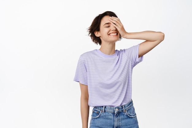 Bella ragazza moderna senza trucco sul viso, pelle pulita e sana, toccando la guancia con gli occhi chiusi e un sorriso sognante, in piedi felice su sfondo bianco