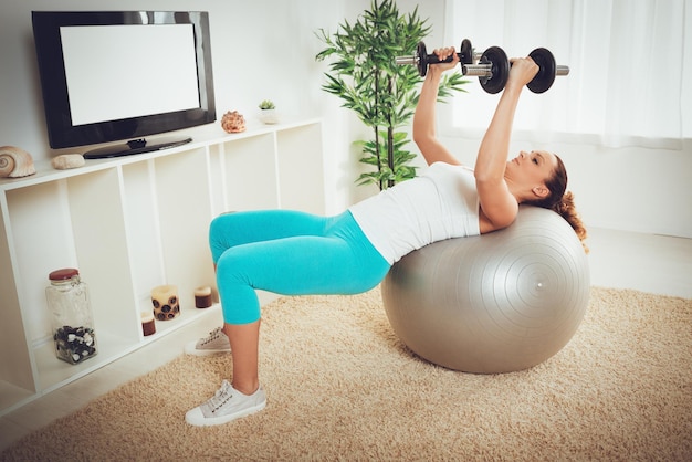 Bella ragazza muscolare che fa allenamento con esercizio con manubri con personal trainer online su una tv a schermo piatto.