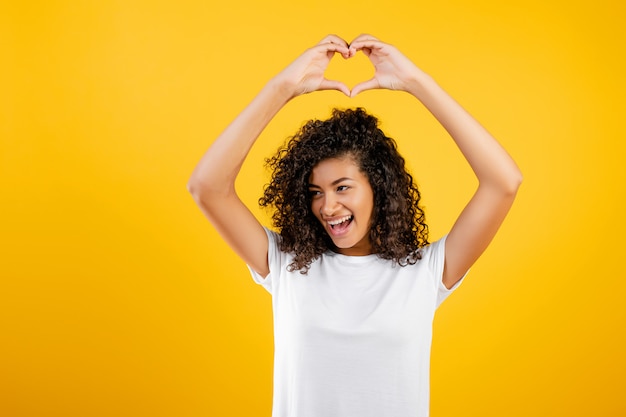 Bella ragazza nera felice che mostra cuore con le mani isolate sopra giallo