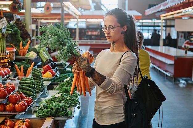 Bella ragazza pensierosa con gli occhiali sta comprando carote fresche al mercato agricolo locale.