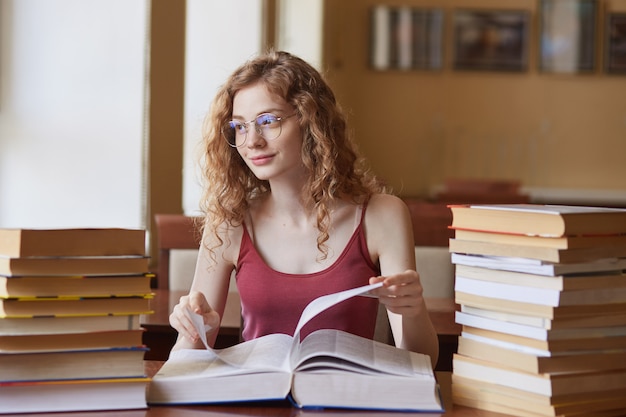 Bella ragazza positiva che ha espressione facciale piacevole, indossando gli occhiali superiori ed alla moda rossi, toccando libro enorme, preparando il suo progetto dello studente all'aula.