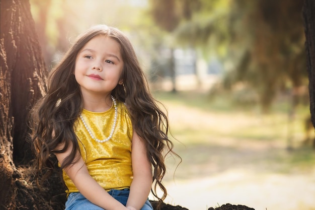 Bella ragazza prescolare bionda seduta su un albero nel tema del giorno dei bambini della ragazza modello della foresta