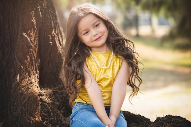 Bella ragazza prescolare bionda seduta su un albero nel tema del giorno dei bambini della ragazza modello della foresta