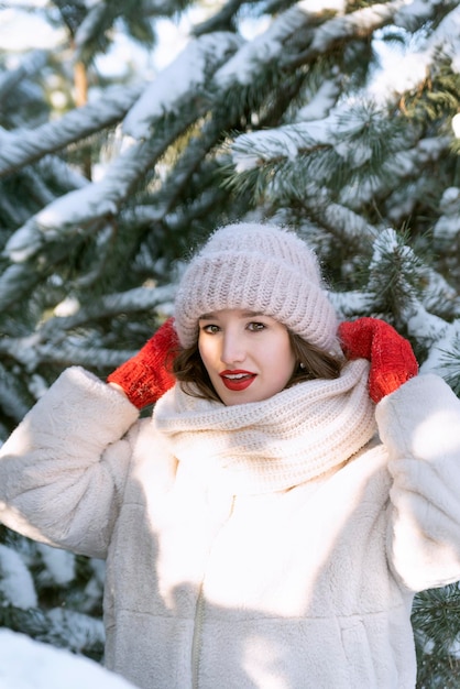 Bella ragazza raddrizza il cappello nella foresta invernale su sfondo di alberi di pino innevato Giornata di sole invernale Cornice verticale