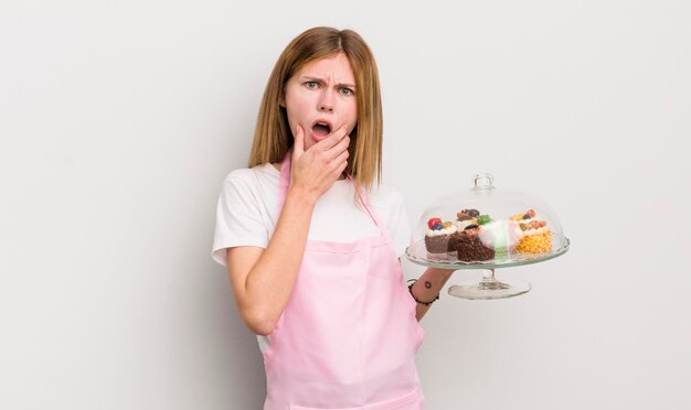 Bella ragazza rossa con la bocca e gli occhi spalancati e la mano sul mento concetto di torte fatte in casa