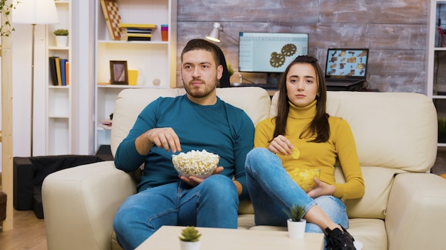 Bella ragazza sdraiata sulla spalla del suo ragazzo mentre guarda la tv e mangia patatine. Fidanzato che mangia popcorn.