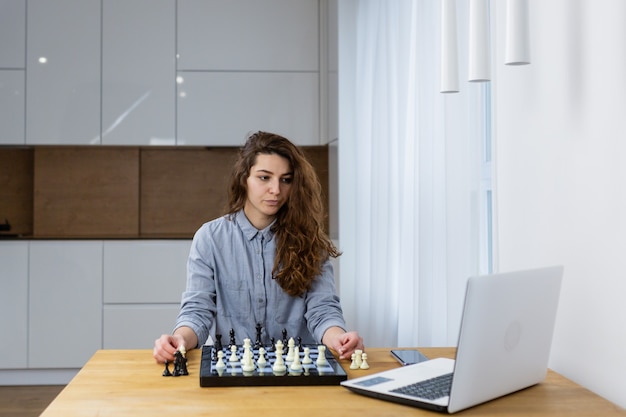 Bella ragazza seduta a casa e giocare a scacchi online con un laptop