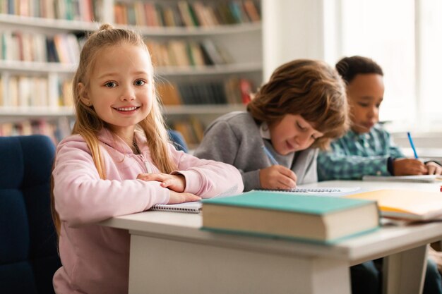 Bella ragazza seduta alla scrivania in classe che sorride alla telecamera studentessa che studia con i compagni di classe dentro