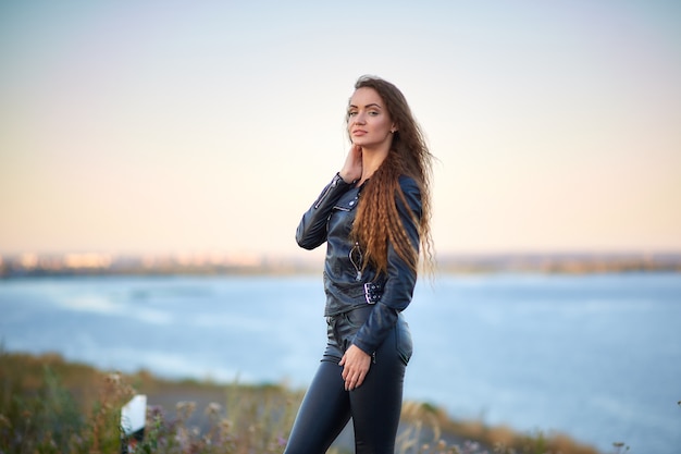 Bella ragazza sexy con i capelli lunghi in una giacca di pelle e pantaloni di pelle in piedi sulla spiaggia al tramonto.