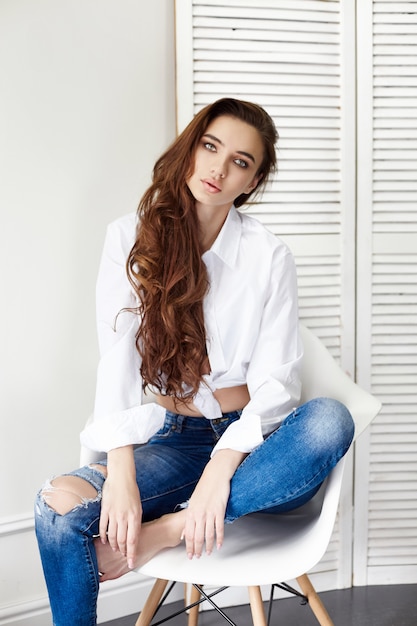 Bella ragazza sexy in camicia bianca dei jeans che si siede su una sedia.