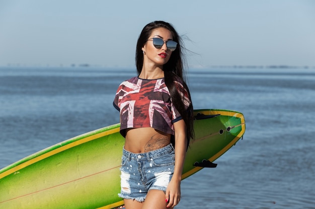 Bella ragazza snella dai capelli scuri in occhiali da sole è in piedi sulla spiaggia sabbiosa vicino al mare e tiene in mano una tavola da surf verde. .