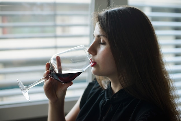Bella ragazza solitaria con bicchiere di vino rosso seduto contro la finestra con gelosia e ammirando il gusto della bevanda alcolica