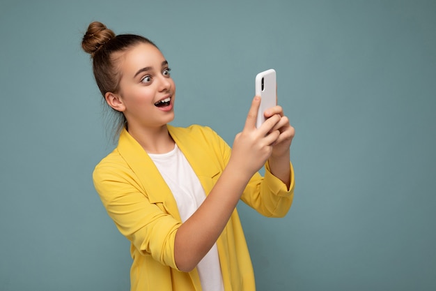 Bella ragazza sorpresa che indossa giacca gialla e t-shirt bianca in piedi isolato su sfondo blu navigando su internet tramite telefono guardando lo schermo del cellulare