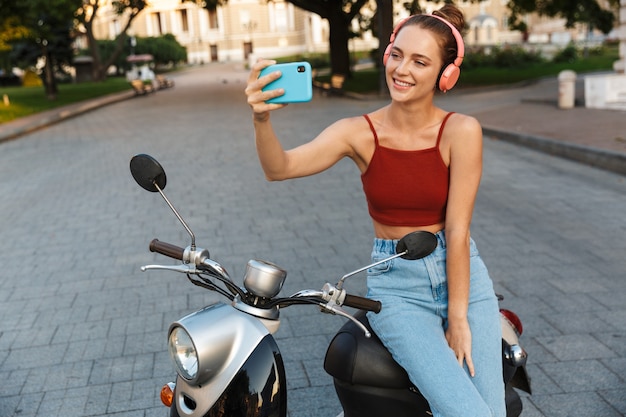 Bella ragazza sorridente che indossa abiti estivi casual seduti su uno scooter all'aperto per le strade della città, facendo un selfie mentre si ascolta musica con le cuffie wireless