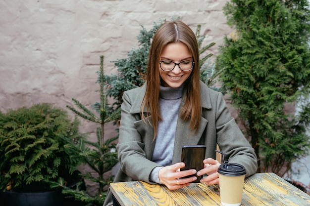 Bella ragazza sorridente con gli occhiali che si siede nella caffetteria tenendo lo smartphone in mano e in chat