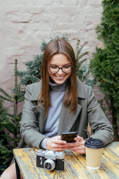 Bella ragazza sorridente con gli occhiali seduta al bar tenendo lo smartphone in mano e chiacchierando