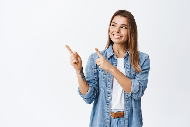 Bella ragazza sorridente con i capelli corti biondi, indicando e guardando a sinistra con un sorriso compiaciuto, facendo la sua scelta per lo shopping, in piedi contro il muro bianco