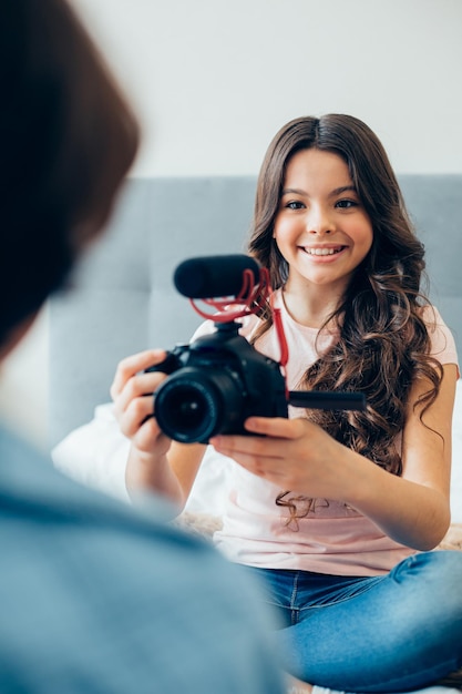 Bella ragazza sorridente e utilizzando una fotocamera moderna con un microfono adattato mentre registra alcuni contenuti per il suo blog