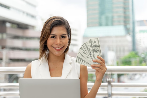 Bella ragazza sorridente in abiti da donna d'affari utilizzando il computer portatile e mostrare denaro Dollaro statunitense in mano sullo sfondo urbano della città