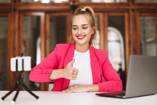 Bella ragazza sorridente in giacca rosa con il laptop che mostra felicemente il pollice in su registrando un nuovo video per il suo vlog