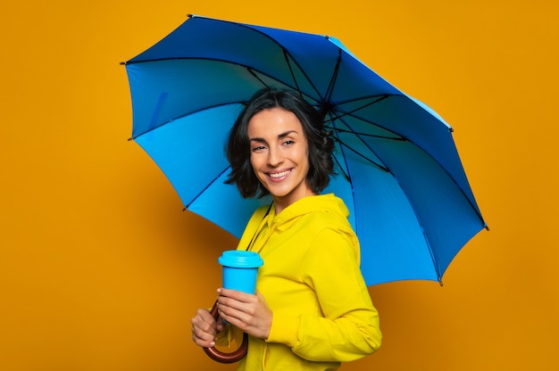 bella ragazza sorridente in una giacca gialla sotto un ombrello, con una tazza termica blu nella mano sinistra, guardando dritto.