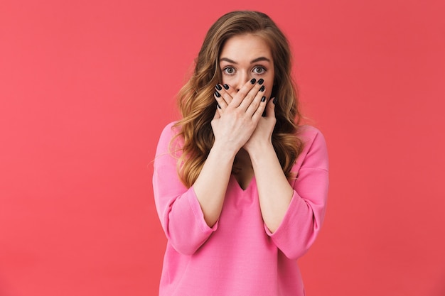 Bella ragazza spaventata che indossa abiti casual in piedi isolata sul muro rosa, copre la bocca