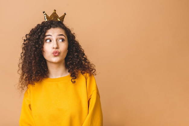 Bella ragazza splendida con capelli marroni ricci e indossare la corona casuale e della tenuta sulla testa isolata sopra il fondo beige dello studio. Invio bacio dell'aria.