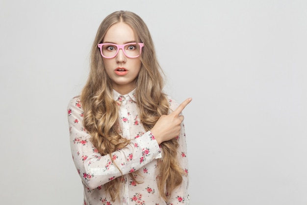 Bella ragazza stressata, che mostra il dito nello spazio della copia e che guarda l'obbiettivo. Studio girato, isolato su sfondo grigio