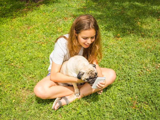 Bella ragazza teenager che guarda un video divertente su uno smartphone con il suo cane in un parco di estate