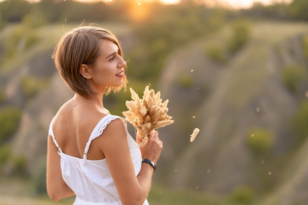 Bella ragazza tenera in un prendisole bianco cammina al tramonto in un campo con un bouquet di spighette.