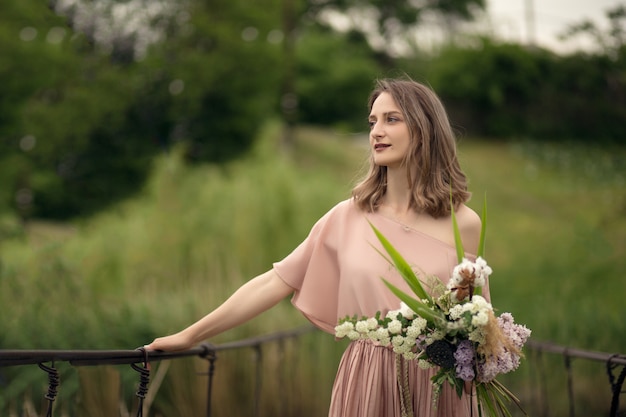 Bella ragazza tenera in un vestito color pesca che cammina su un ponte con un mazzo di fiori