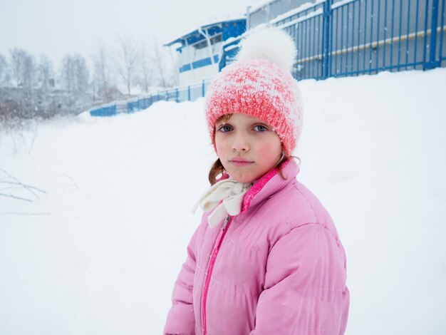 Bella ragazza triste di anni su uno sfondo di neve, un cappello lavorato a maglia rosa e un inverno rosa caldo