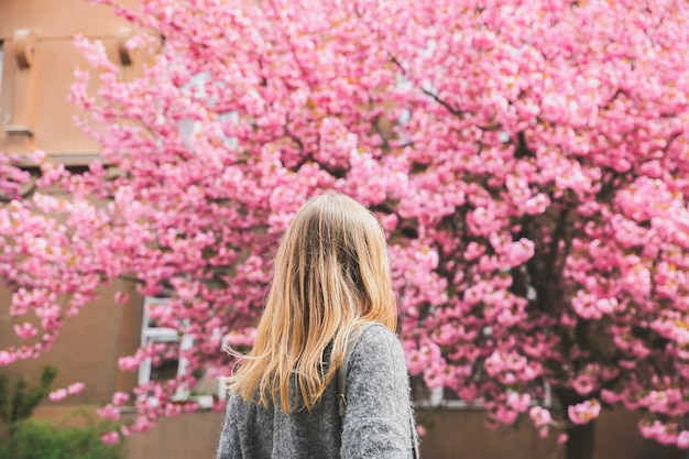 Bella ragazza vicino agli alberi di sakura. Donna in abito e cappotto elegante Fiori rosa che sbocciano a Uzhhorod. Sbocciare intorno. Tempo di primavera. Concetto di relax e felicità.