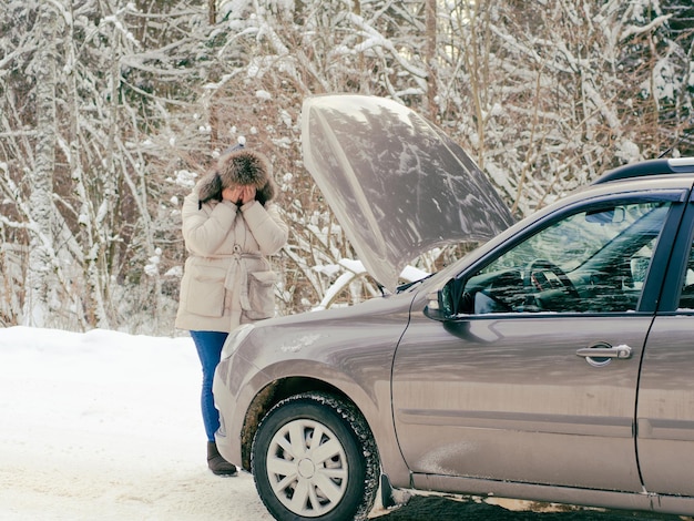 Bella ragazza vicino al motore di un'auto rotta su una strada innevata d'inverno. si è bloccato e chiede aiuto