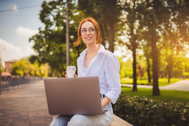 Bella rossa giovane donna seduta nel parco e utilizzando laptop studente universitario freelance wear...