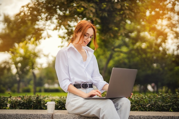 Bella rossa giovane donna seduta nel parco e utilizzando laptop studente universitario freelance wear...