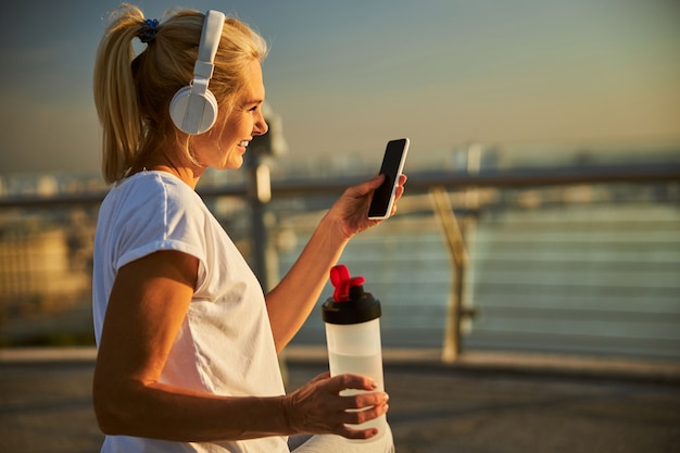 Bella signora che tiene in mano una bottiglia d'acqua e sorride mentre ascolta la musica tramite le cuffie wireless e utilizza lo smartphone