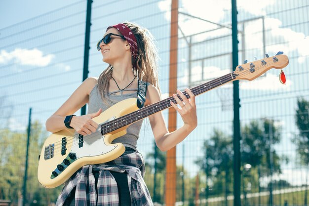 Bella signora con i dreadlocks che indossa occhiali da sole e sorride mentre guarda in lontananza e suona la chitarra al campo sportivo