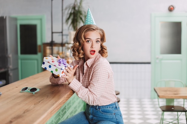 Bella signora con il cappello di compleanno seduta al bancone del bar con la scatola del regalo in mano e guardando sorprendentemente a porte chiuse nella caffetteria