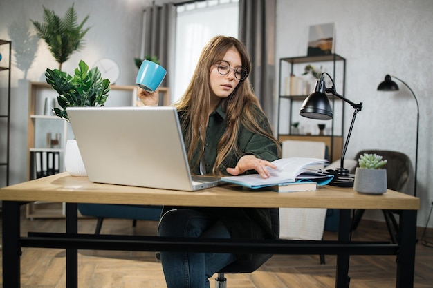 Bella signora con note di lettura di bevande calde mentre si lavora al computer portatile