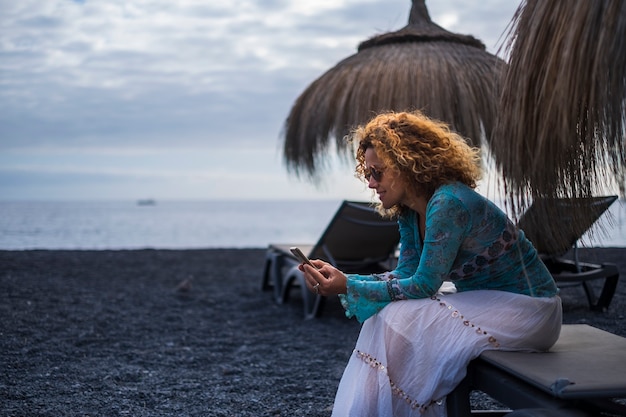 bella signora di mezza età che utilizza lo smartphone tecnologico in spiaggia durante le vacanze invernali da solo. vestito casual hippy chic e sorriso. oceano. toni blu. concetto di indipendenza