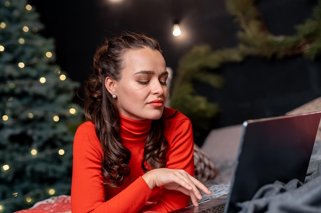 Bella signora in abiti rossi sdraiata davanti al suo laptop Donna con il taccuino sul plaid vicino all'albero di Natale Donna con i capelli ricci che lavora al suo laptop vicino all'albero di abete