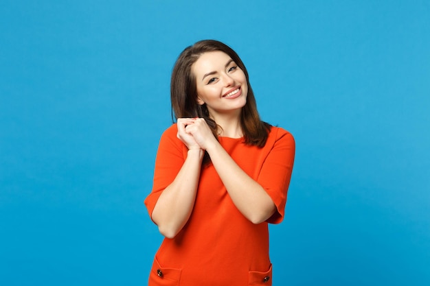 Bella sognante giovane donna bruna che indossa un abito rosso arancio che fa il desiderio in piedi in posa isolata su sfondo blu muro, ritratto in studio. Concetto di moda stile di vita delle persone. Mock up spazio di copia.