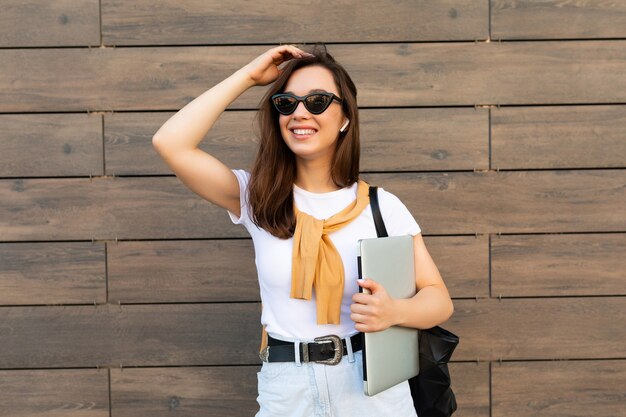 Bella sorridente affascinante giovane donna brunet che guarda l'obbiettivo tenendo il computer portatile e occhiali da sole in t-shirt bianca e jeans azzurri in strada.