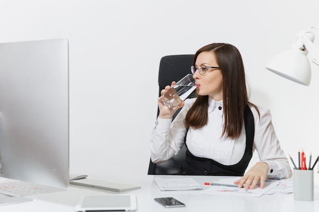 Bella sorridente donna d'affari dai capelli castani in giacca e occhiali seduta alla scrivania, bevendo acqua pura, lavorando al computer con monitor moderno con documenti in ufficio leggero