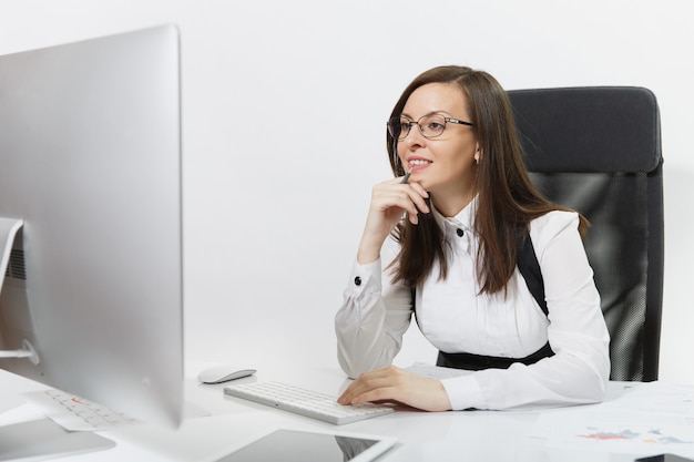 Bella sorridente donna d'affari dai capelli castani in giacca e occhiali seduta alla scrivania, che lavora al computer con un monitor moderno con documenti in ufficio leggero